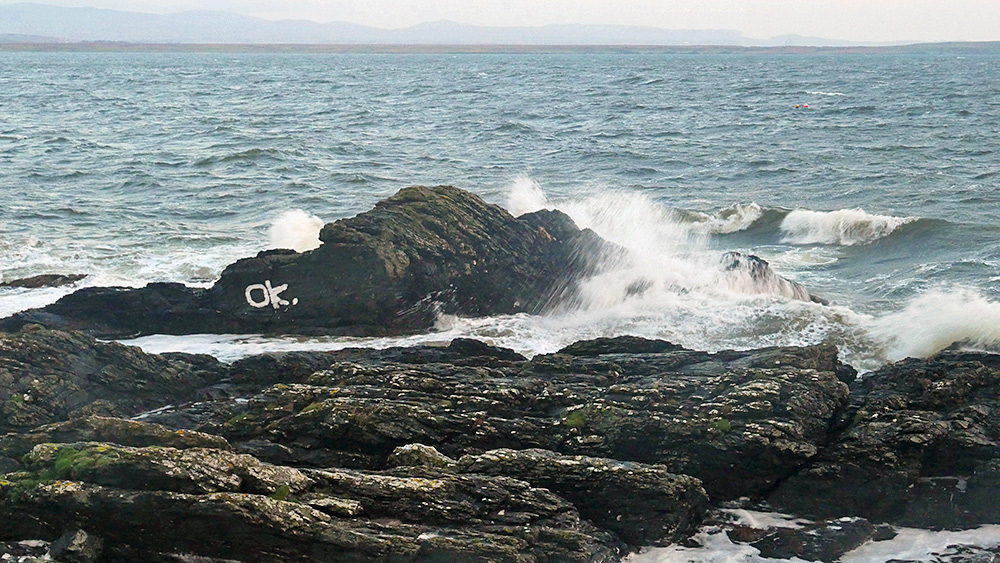 Picture of a small rock with the letters OK painted on it just off a rocky shore, waves splashing over the rock