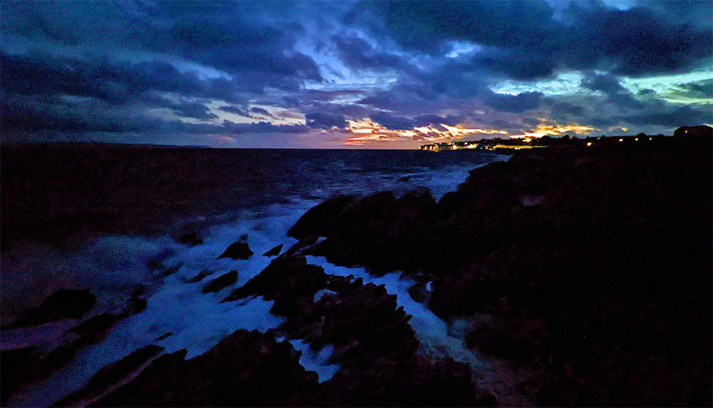 Picture of a rocky shore with low cliffs and breaking waves, a coastal village in the distance in the last light of the gloaming