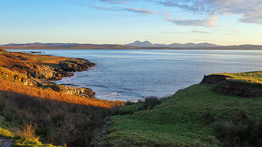 Picture of a sea loch shoreline in the mild morning sunshine, mountains visible in the distance across the loch