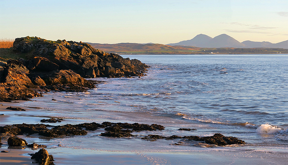 Picture of a small beach with some coastal rocks at a sea loch, mountains in the distance across the loch