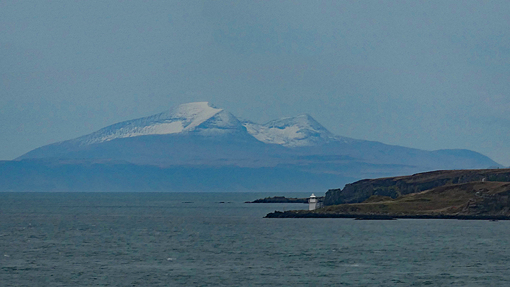 Picture of a snow capped mountain seen across the sea from a sound between two islands, a small lighthouse on the shore