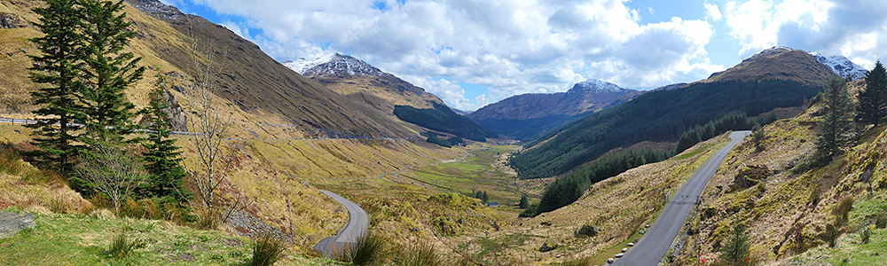 Panoramic picture of a view down a glen/valley from a viewpoint at the top of it, some mountains with snow on the tops around