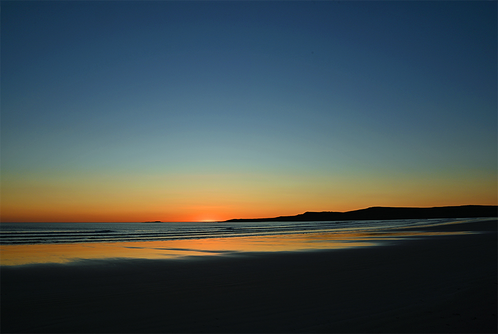 Picture of the last seconds of an April sunset with the sun just disappearing behind the horizon, seen from a wide sandy beach