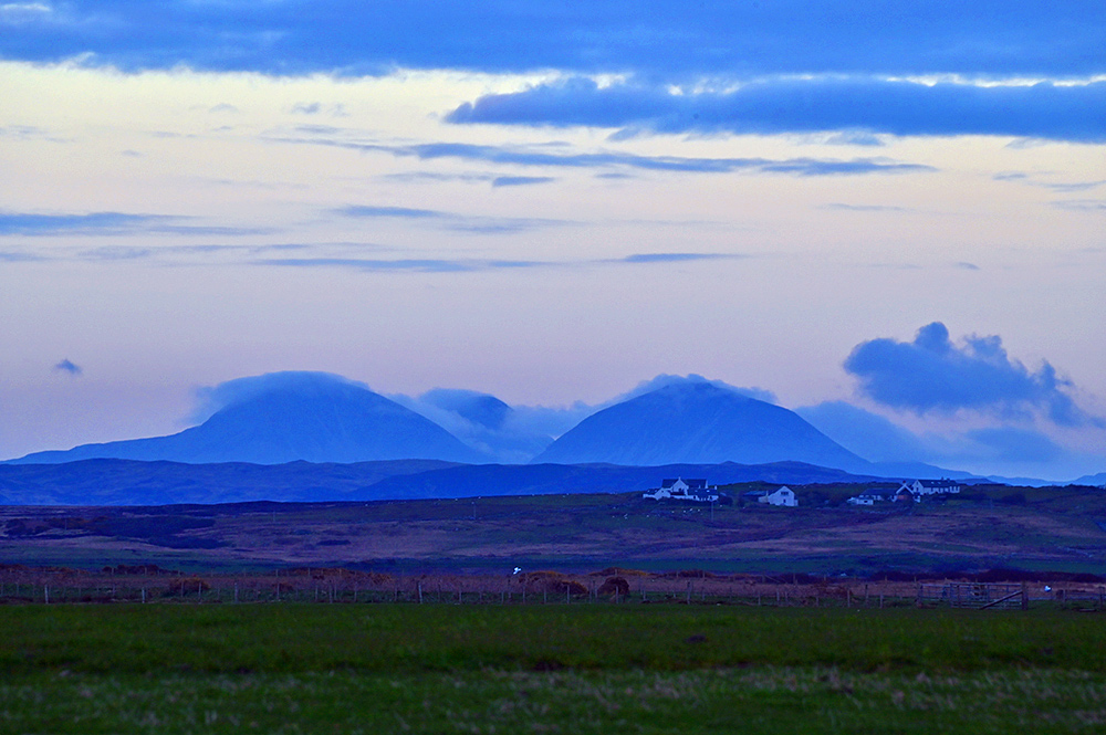 Picture of three mountains with low clouds hanging around them in the failing evening light, a small settlement in the foreground
