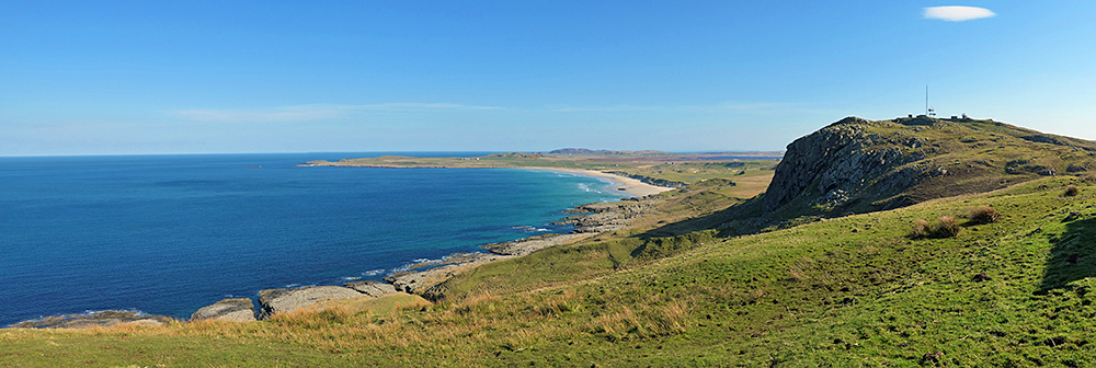 Panoramic picture of a wide bay with a beach, crags with an old radar station on top next to it