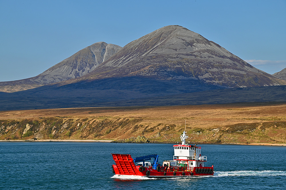 Picture of a small red landing craft in a sound between two islands, two mountains in the background