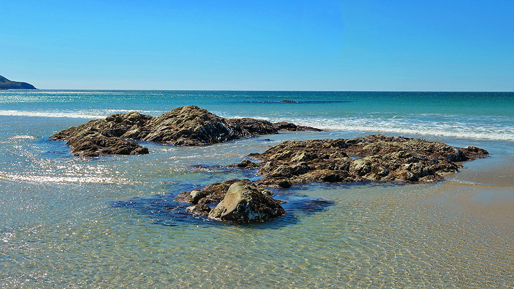 Picture of some rocks in shallow water near the end of a beach on a bright sunny day