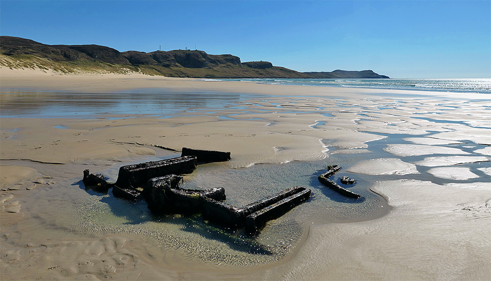 Picture of the remains of an old wreck embedded in a sandy beach at low tide under a bright sunny blue sky