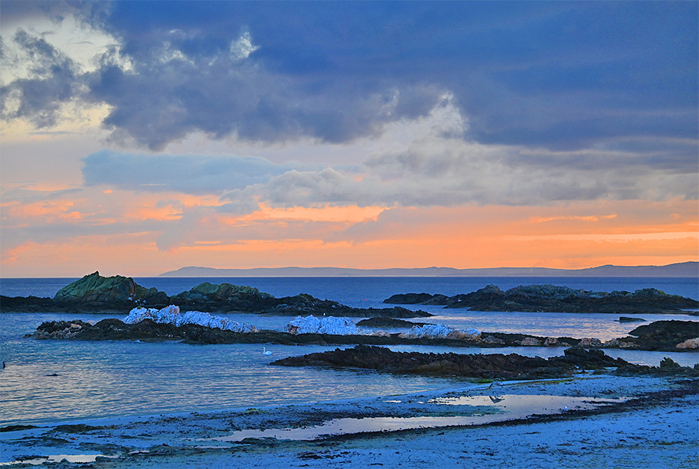 Picture of a beach with rocky outcrops at an approaching sunset, another island in the distance