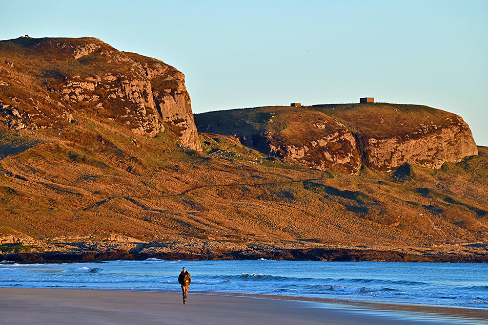 Picture of a walker on a beach in the evening sunset light under steep crags