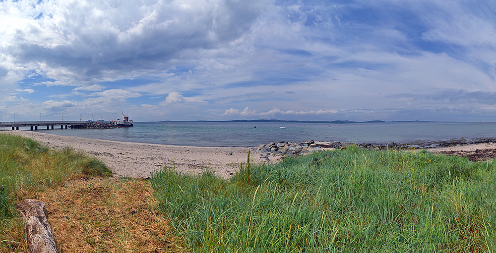 Panoramic picture of a small pier with a small ferry and island in the distance, seen from a beach