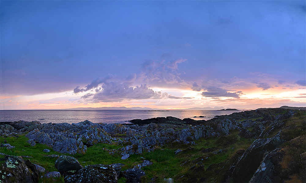 Panoramic picture of a view from a rocky shore over some water to some islands during a cloudy sunset