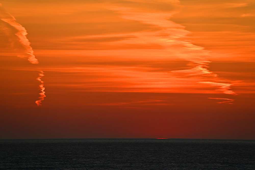 Picture of the very end of a sunset with just a tiny sliver of light visible at the horizon under a colourful red sky