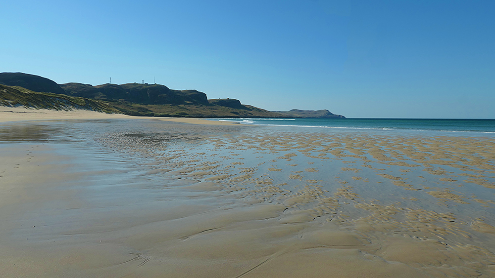 Picture of a beach with shallow puddles on the sand, steep crags in the distance under a clear blue sky