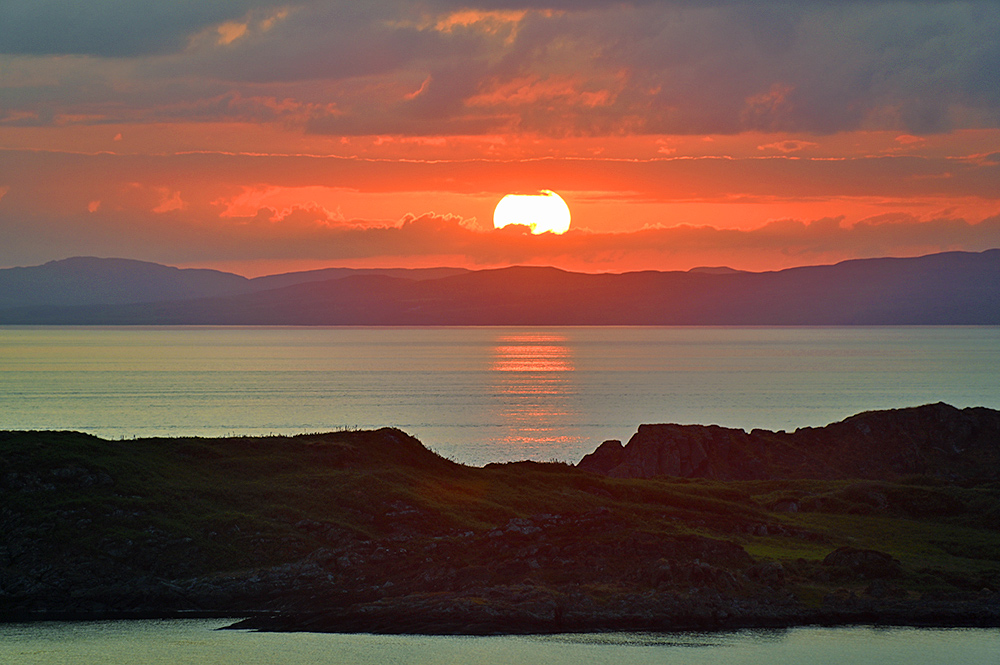 Picture of a cloudy sunset with the sun briefly breaking through the clouds over two islands, seen from a third island
