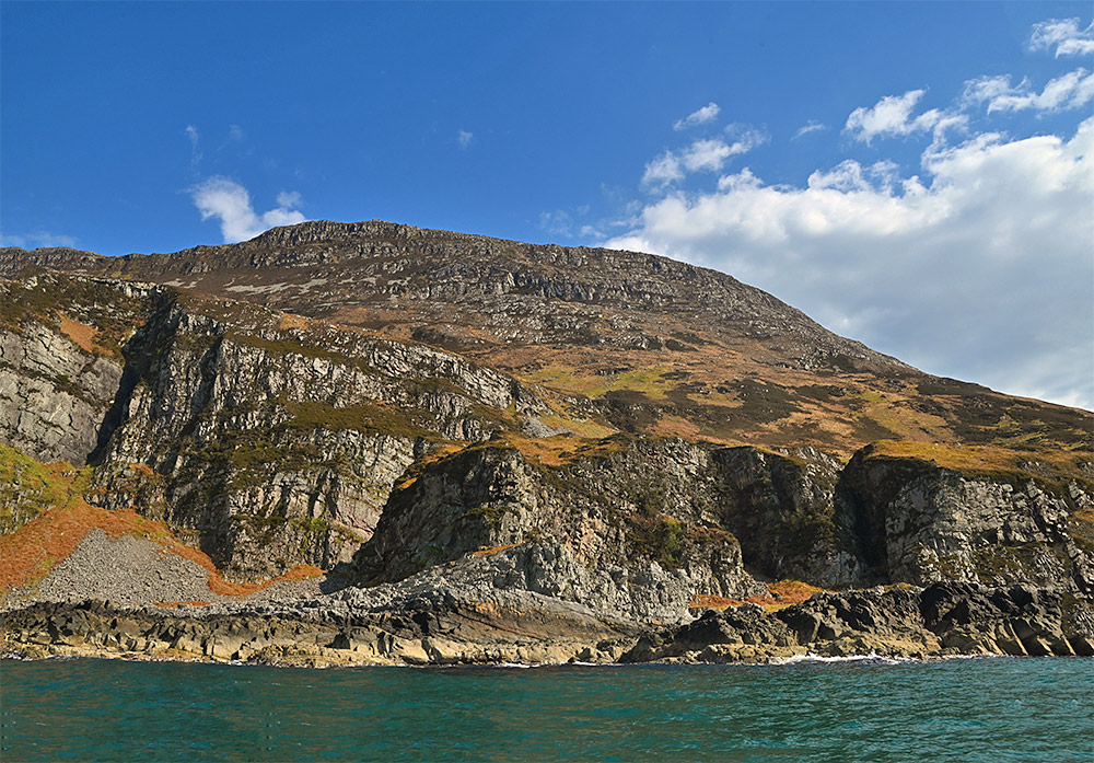Picture of steep cliffs above a rocky shore seen from a boat on the water