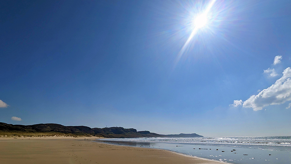 Picture of a bright sun in a blue sky over a bay with a wide sandy beach with crags and cliffs in the distance