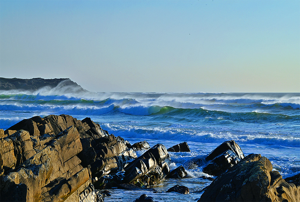 Picture of a bay with a rocky shore, waves rolling in and spray blown back by the wind