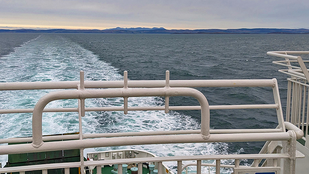 Picture of some higher hills behind a lower coastline, seen from the stern of a ferry