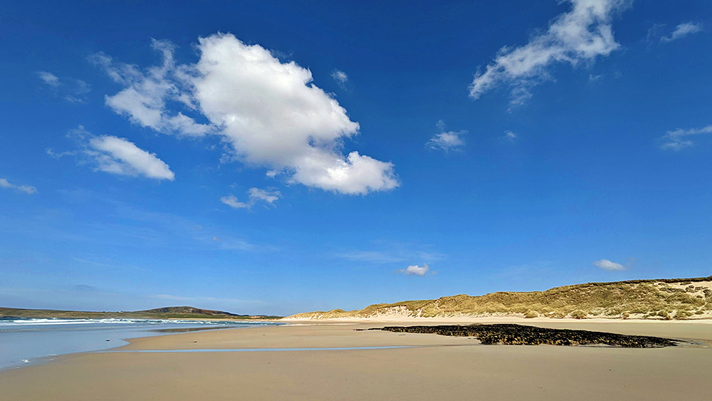 Picture of a big fluffy cloud (and a few smaller clouds) over a wide sandy beach with dunes at the back