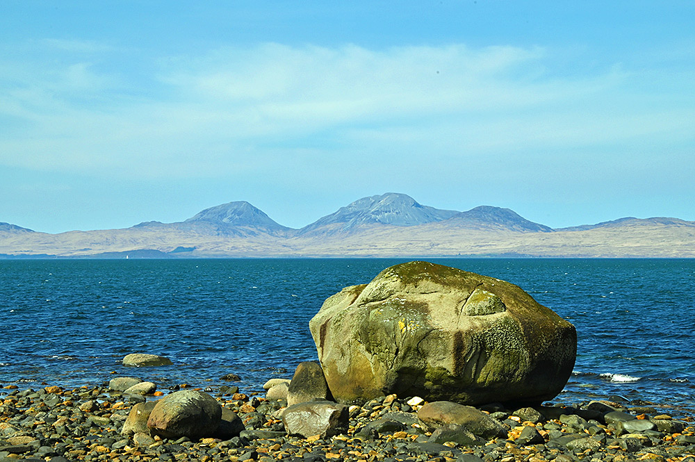 Picture of a very large boulder at a stone beach, three mountains in the distance behind