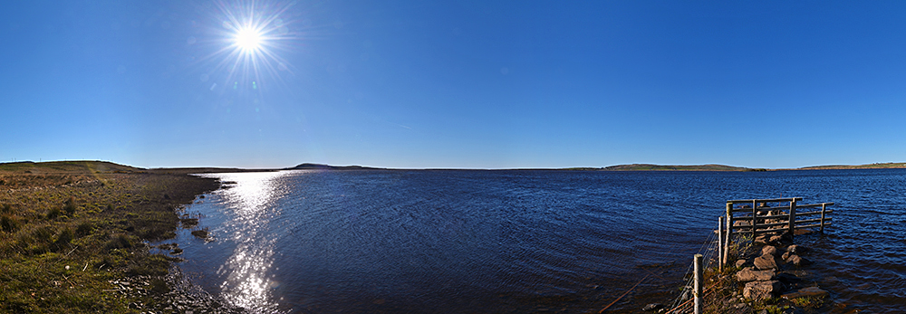 Panoramic picture of a bright sun in a blue sky over a large freshwater loch