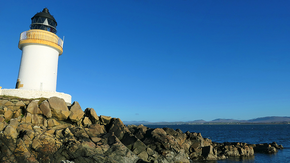 Picture of a small lighthouse on a rocky shore of a sea loch, a village visible in the distance on the other side of the loch