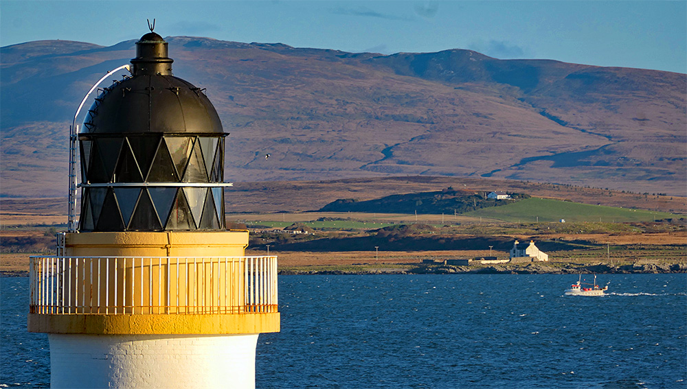 Picture of the top of a lighthouse, behind it a sea loch with an old farm house at the shore and a passing small fishing boat