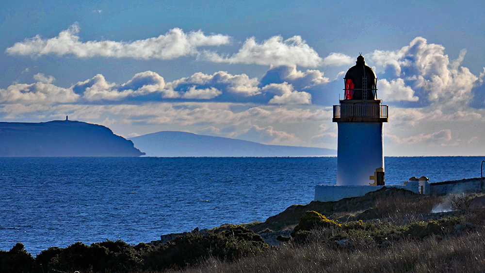 Picture of a small coastal lighthouse with the mull of a peninsula in the distance. Another mountain in the far distance