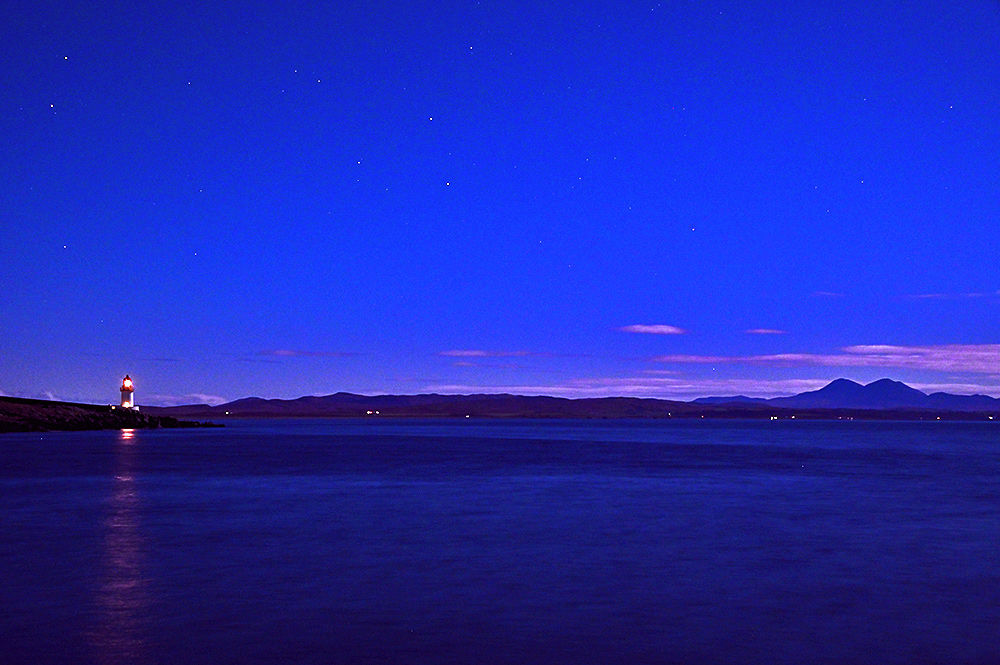 Picture of a sea loch with a small lighthouse at night, some mountains visible in the distance