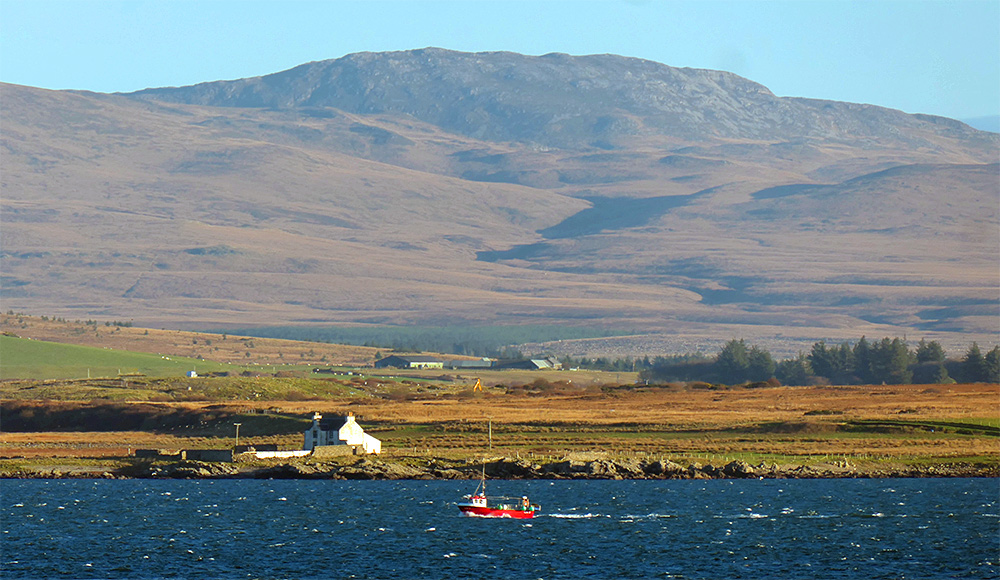 Picture of a red fishing boat passing an old farm house near the shore of a sea loch