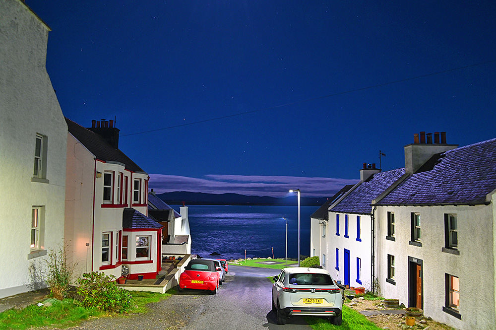 Picture of a village street leading down to a sea loch at night, some stars visible in the sky