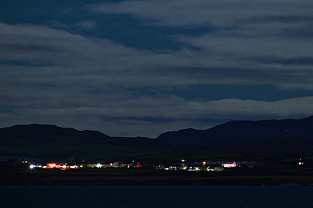 Picture of a coastal village at night seen across a sea loch, only the lights visible below dark hills