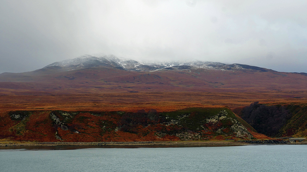 Picture of some low hills with a dusting of snow under low clouds, seen from a ferry in a sound between two islands