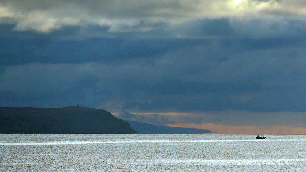 Picture of a fishing boat off a mull at the end of a peninsula with a monument on top