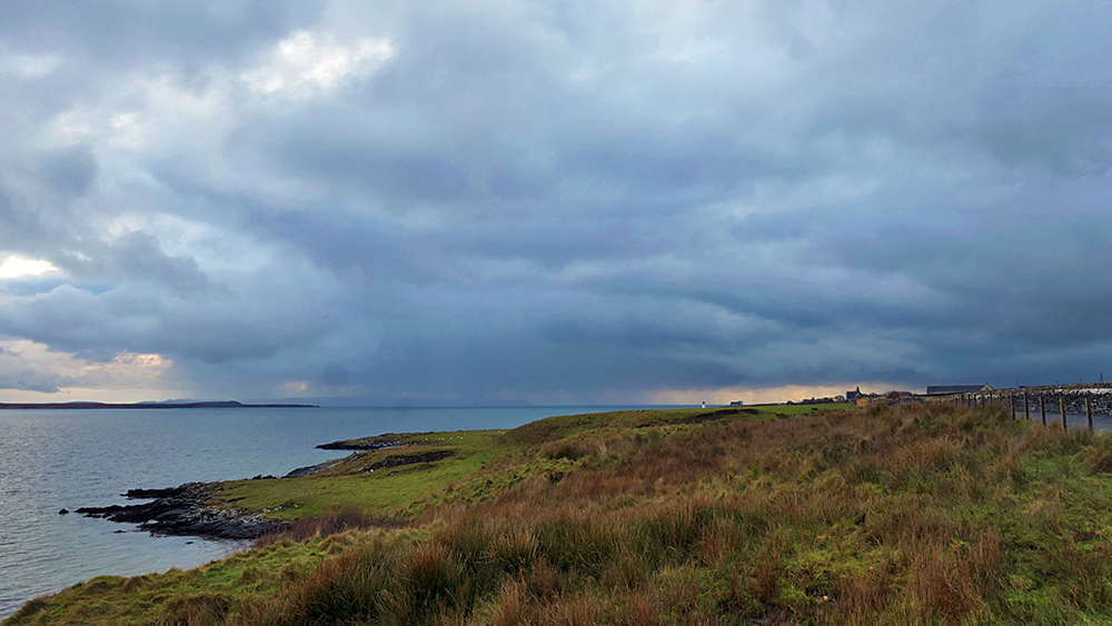 Picture of a view over a sea loch with a grassy shoreline on a cloudy day, some heavy rain approaching in the distance