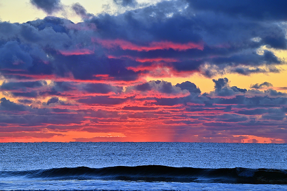 Picture of some colourful mostly red clouds in the gloaming after sunset over a single breaking wave in a wide bay