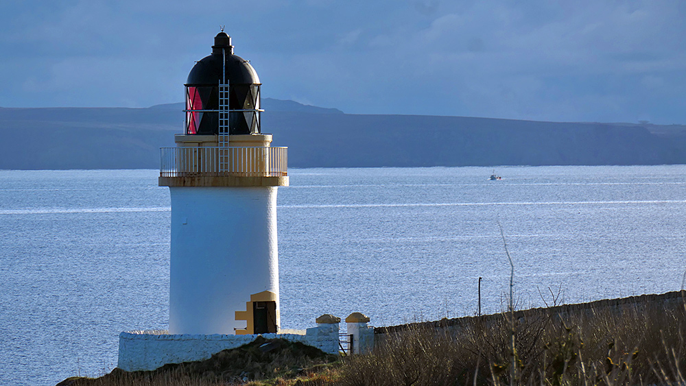 Picture of a small lighthouse on the shore of a sea loch, a fishing boat approaching in the distance
