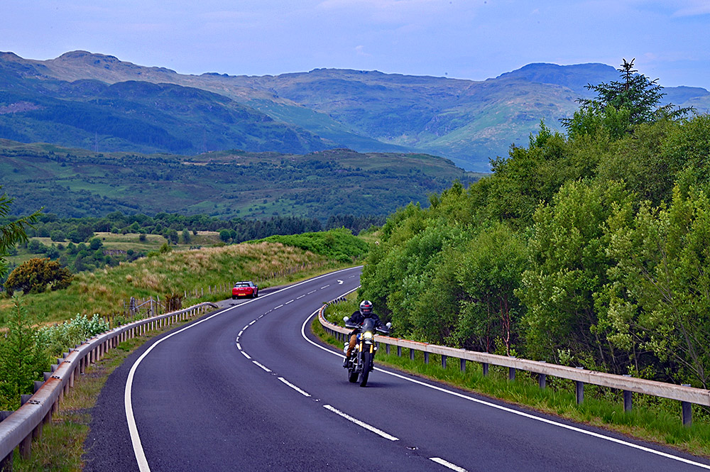 Picture of a road through hills, a motorbike passing. A red car parked in a layby in the distance