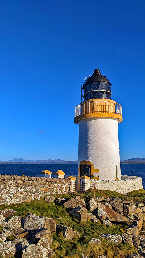 Picture of a small white lighthouse on a sea loch shore on a sunny day, some mountains in the distance