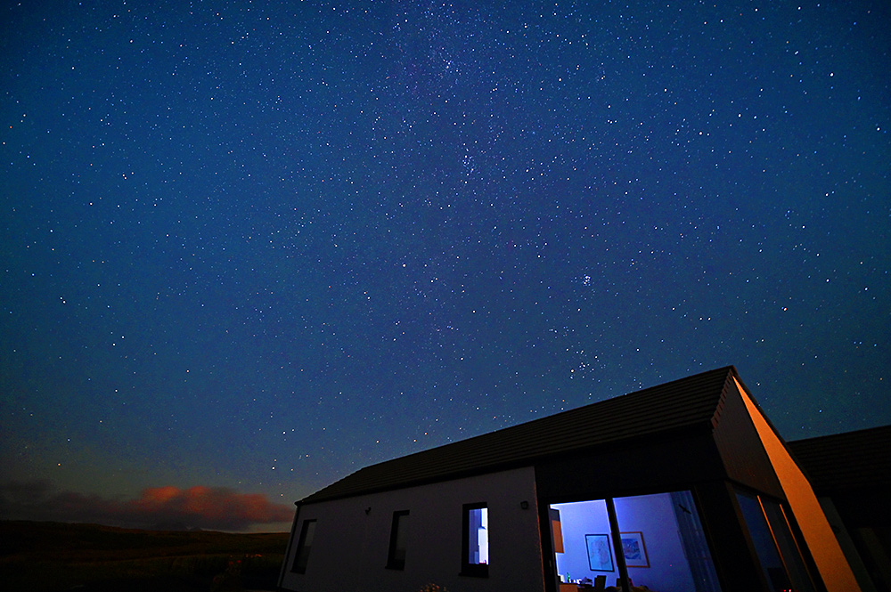 Picture of a clear starry night sky above a house with some light inside, some clouds on the horizon