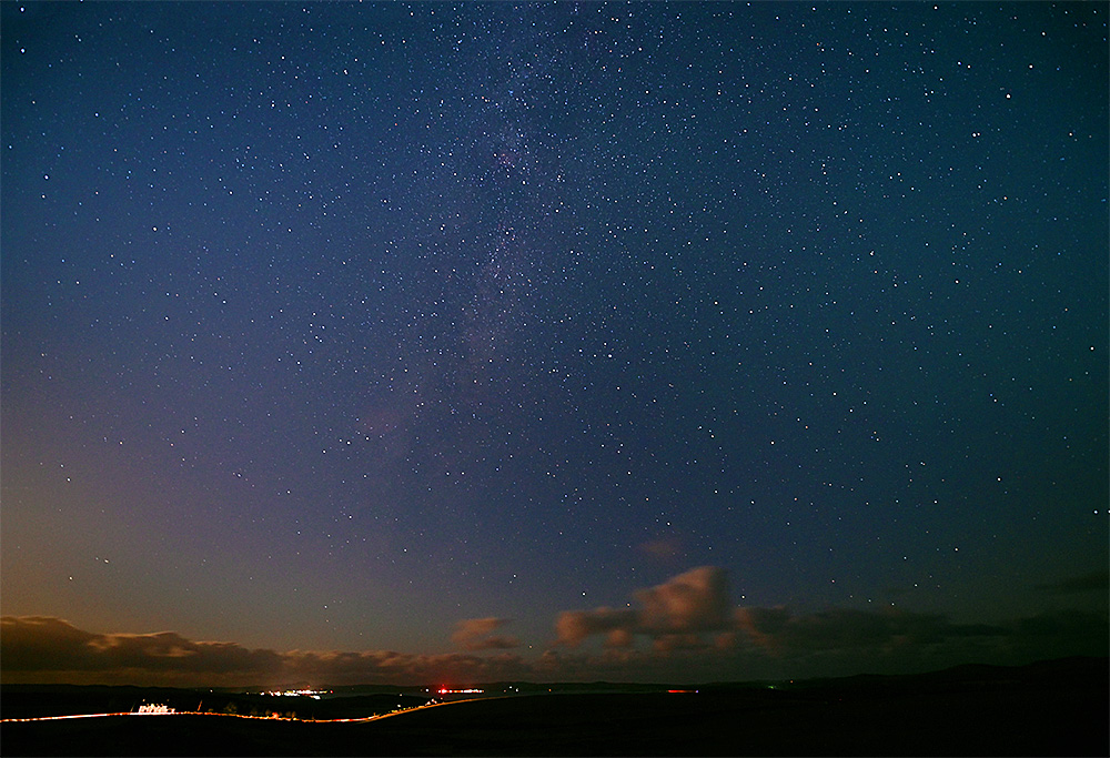 Picture of a wide starry night sky seen from a hill. Two villages in the distance, a road lit up by a passing car below