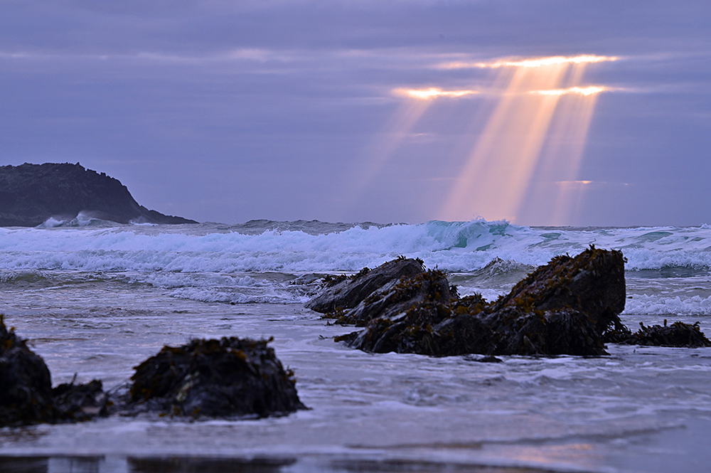 Picture of some sunrays breaking through clouds over a bay with rocks in a beach and waves rolling in