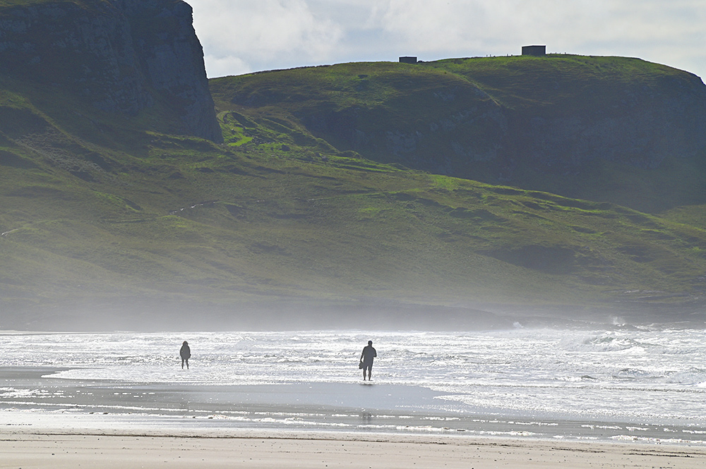 Picture of two beach walkers waking barefoot in the shallow surf of a bay with some crags in the background