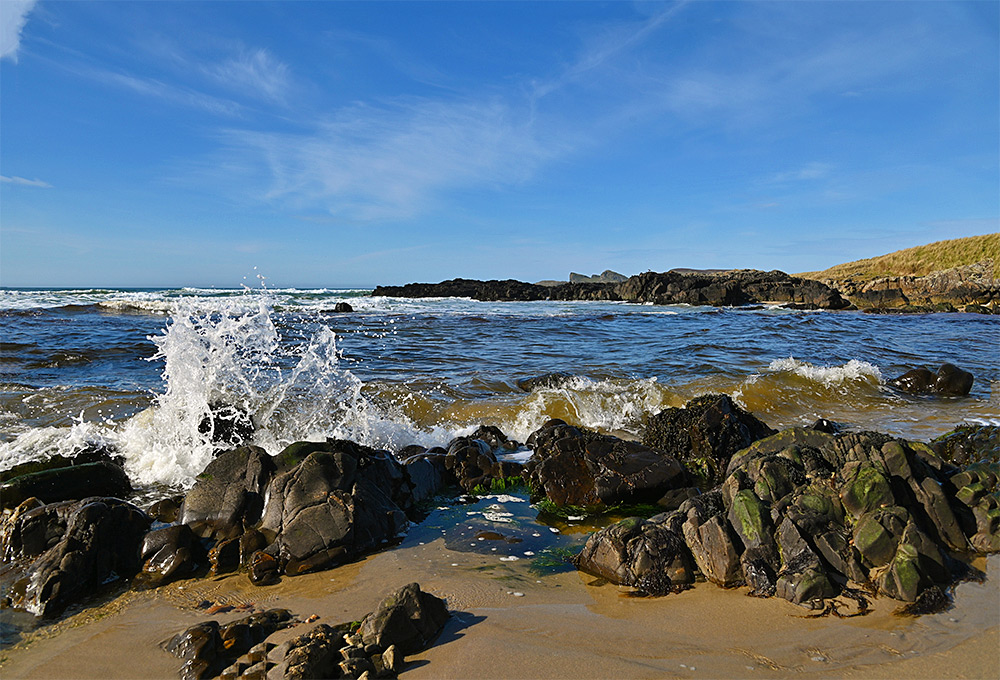 Picture of a small wave splashing over some rocks on a sandy beach, rocks and dunes in the background, a rock formation in the distance