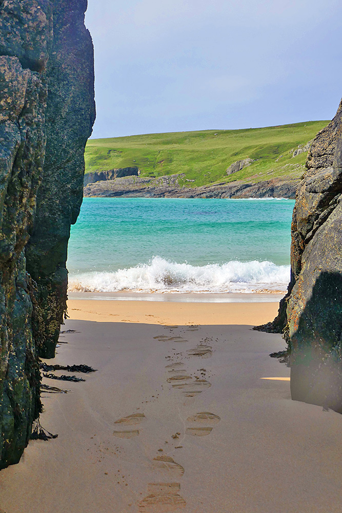 Picture of some footprints in the beach sand between some rocks, a wave breaking on the beach in the background