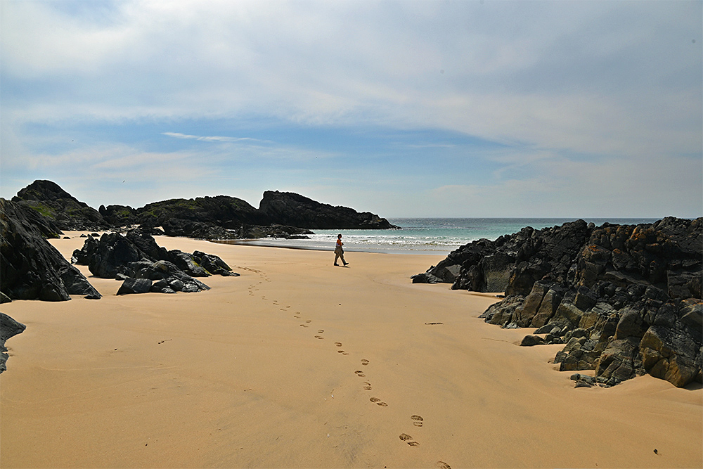 Picture of a woman walking on a golden sandy beach with some dark rocks