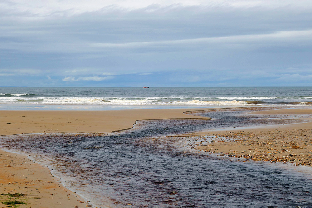Picture of a small river flowing over a beach into the sea. A small fishing boat visible in the distance.