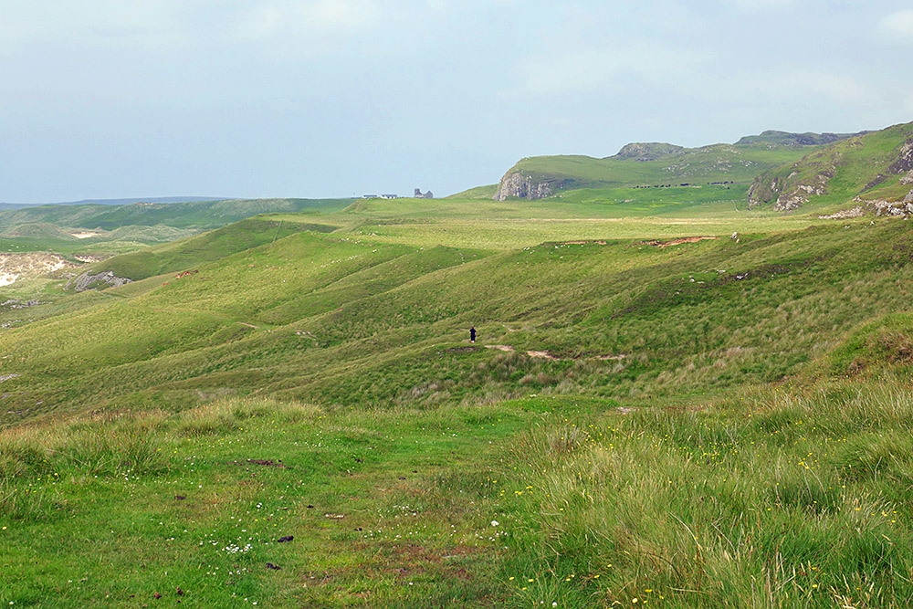 Picture of a rough track along a hillside, some crags on the side and a church ruin in the distance. A person just visible in the distance walking down on the track