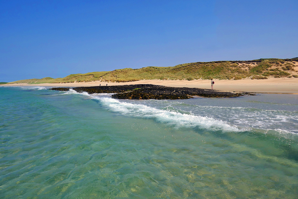 Picture of a rocky outcrop on a sandy beach with dunes, seen from the water